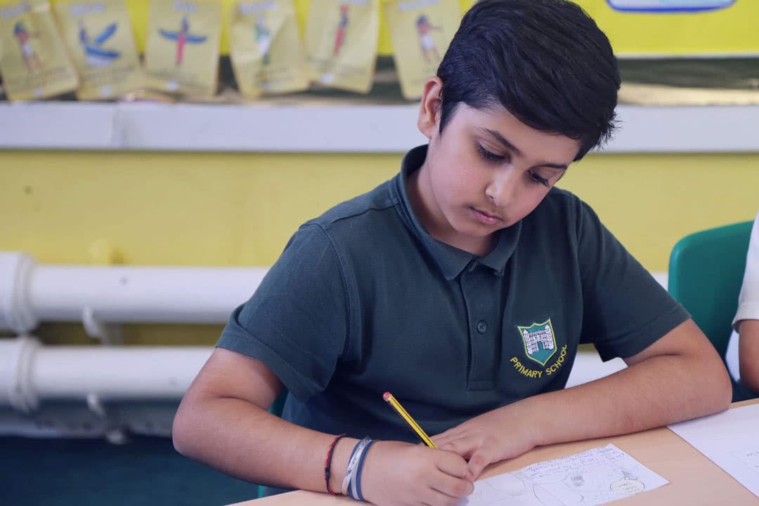 A primary school boy in green uniform is sat in his classroom writing with a pencil.