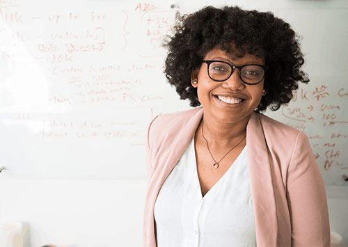 A teacher wearing glasses is smiling, standing in front of a whiteboard with mathematically equation on.